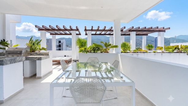 Un patio moderno al aire libre con mesa y sillas de vidrio, una parrilla y una pérgola con plantas.