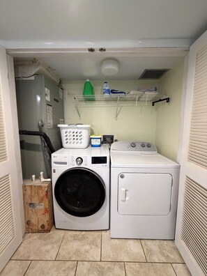 A laundry room with two washing machines, a drying machine, a shelf with cleaning supplies, and a vent. - #207 3BR 3.5BA  Direct Ocean, Full Remodel, 2K + 2Q, Indoor Laundry, Gorgeous (Daytona Beach Shores)