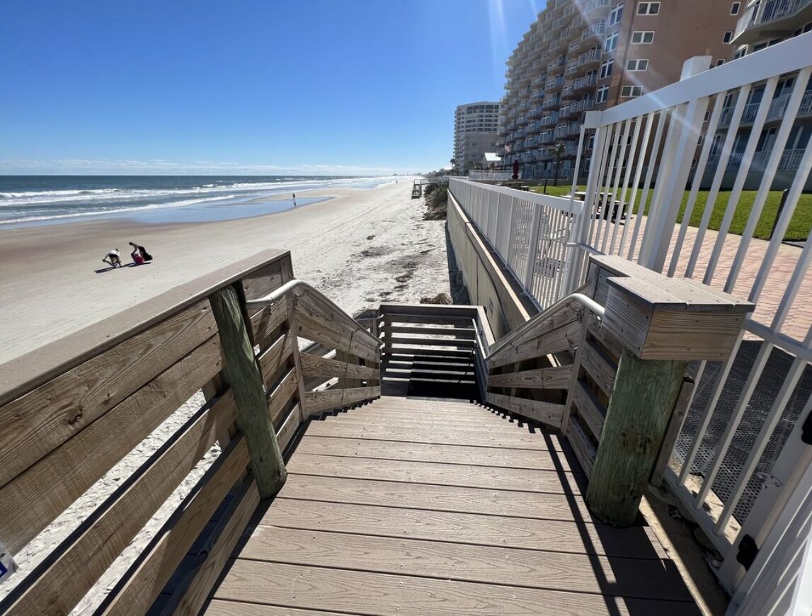 Una escalera de madera que lleva a una playa, con vista al océano y un edificio de gran altura al fondo.
