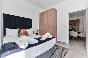 Quarto de hotel com cama, parede de destaque em madeira, área de refeições com cadeiras e cozinha visível através de uma porta aberta.
