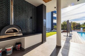 Un espace cuisine extérieur doté d’un four à pizza, de pots de peinture et offrant une vue sur une piscine et sur la verdure.
