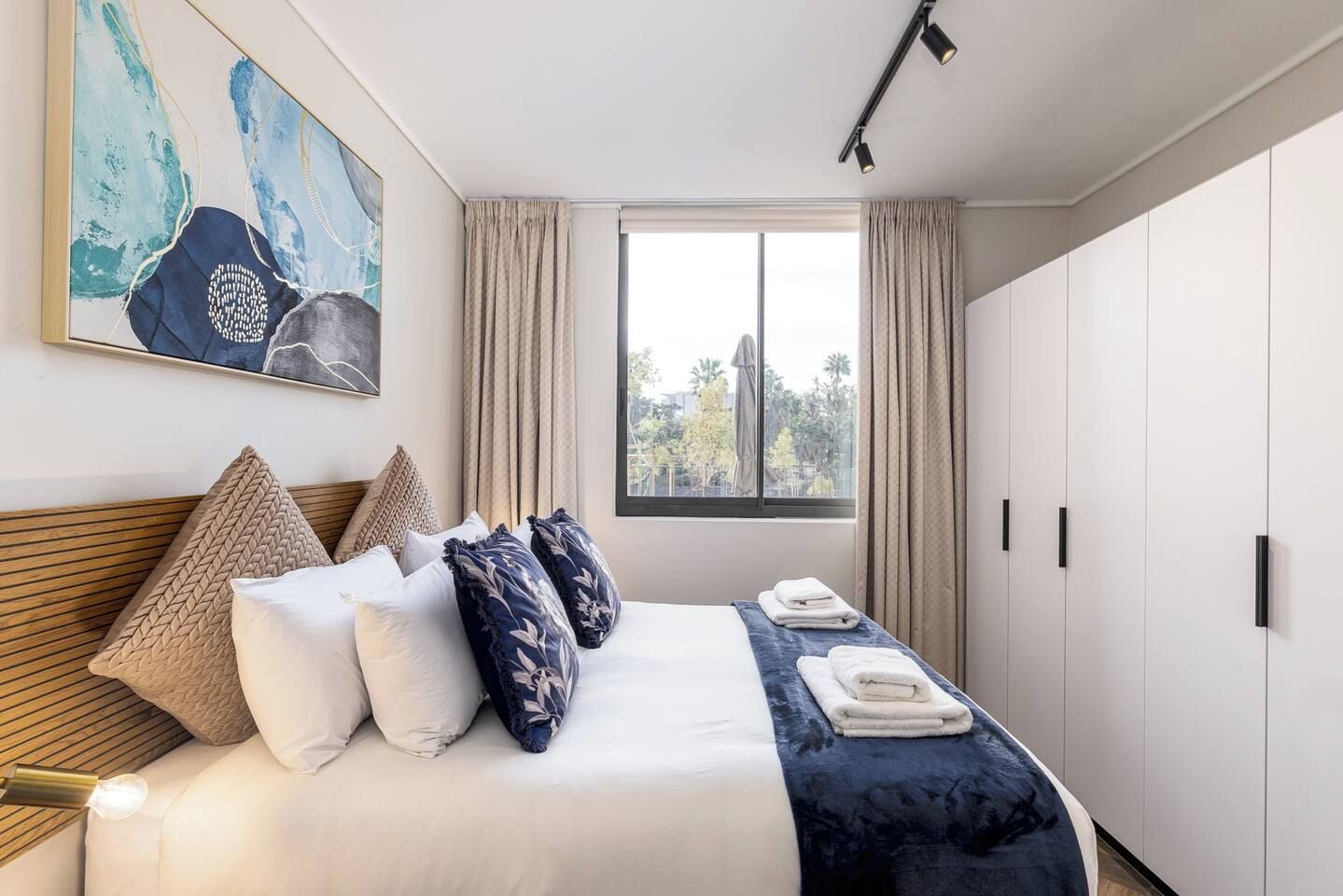 A hotel room with a bed, a window, a wardrobe, and a painting on the wall.