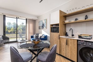 A modern kitchen with a built-in washing machine, a dining area with a glass table and chairs, and a view of the outdoors through large windows. - Modern Luxury at Park Place by Ctha (Cape Town)