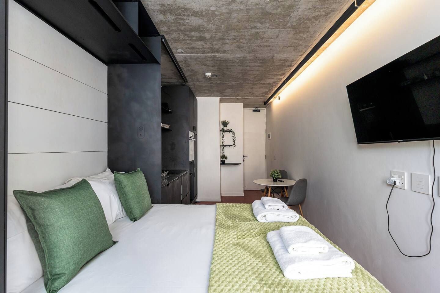 A modern hotel room with a bed, a flat-screen TV, a small dining table, and a kitchenette.