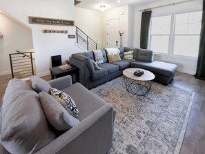 Ein modernes Wohnzimmer mit einem grauen Ecksofa, einem runden Couchtisch und einer Treppe mit Metallgeländer.