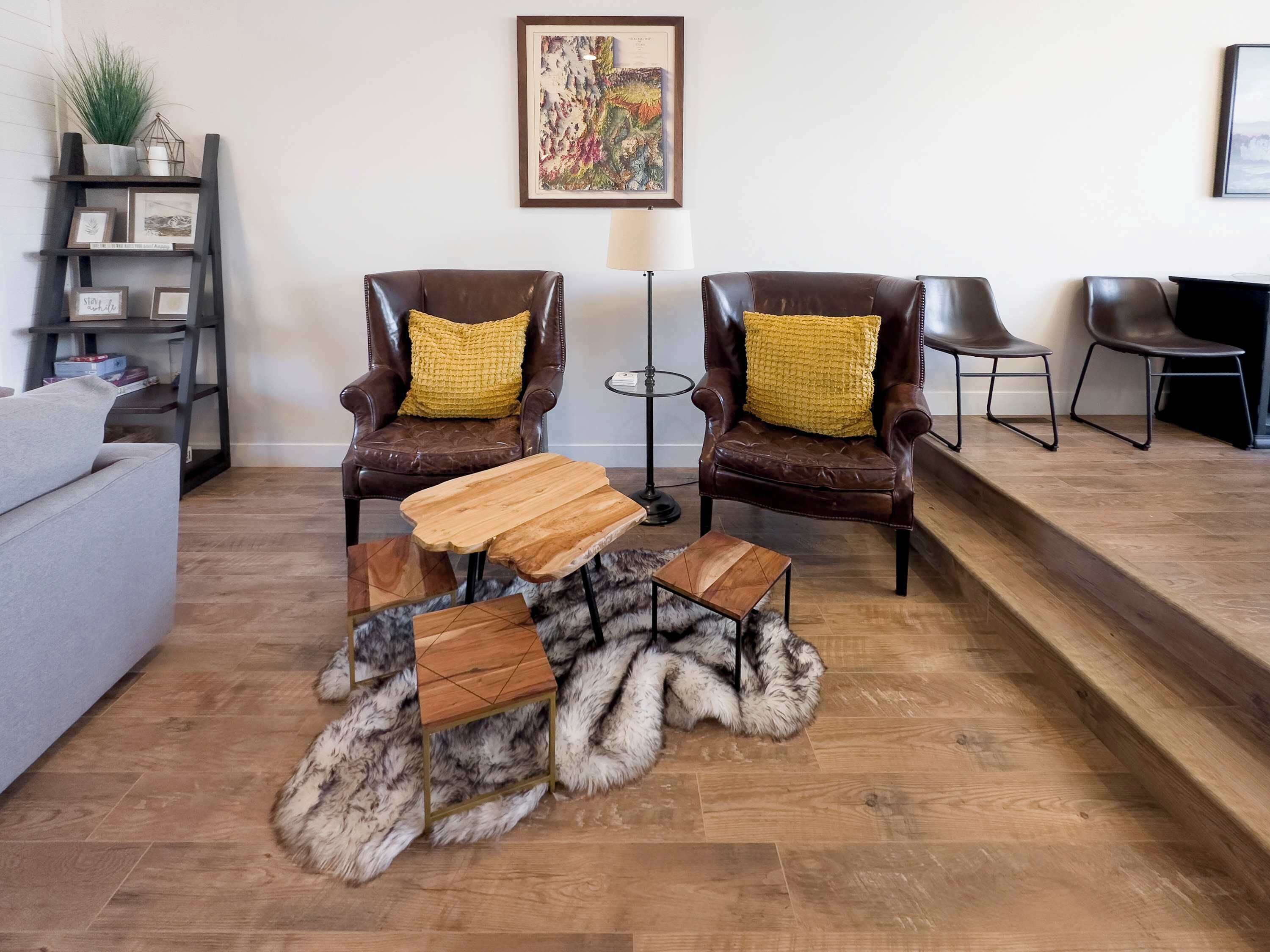 A living room with two leather armchairs, a wooden coffee table, a fur rug, a grey sofa, a bookshelf, and a framed artwork on the wall.
