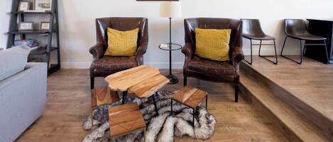 A living room with two leather armchairs, a wooden coffee table, a fur rug, a grey sofa, a bookshelf, and a framed artwork on the wall.