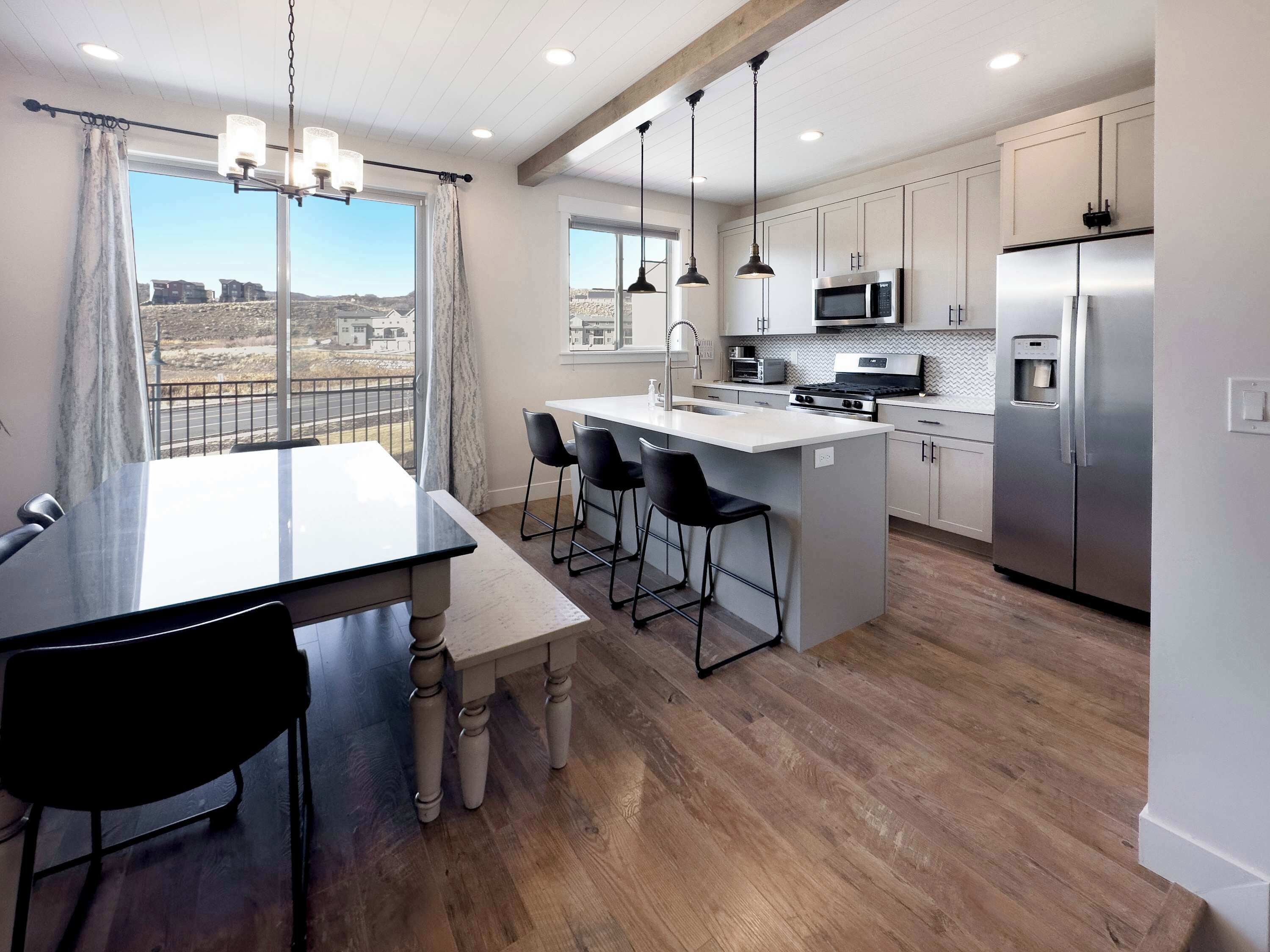 A modern kitchen with a central island, bar stools, stainless steel appliances, and a view through large windows.