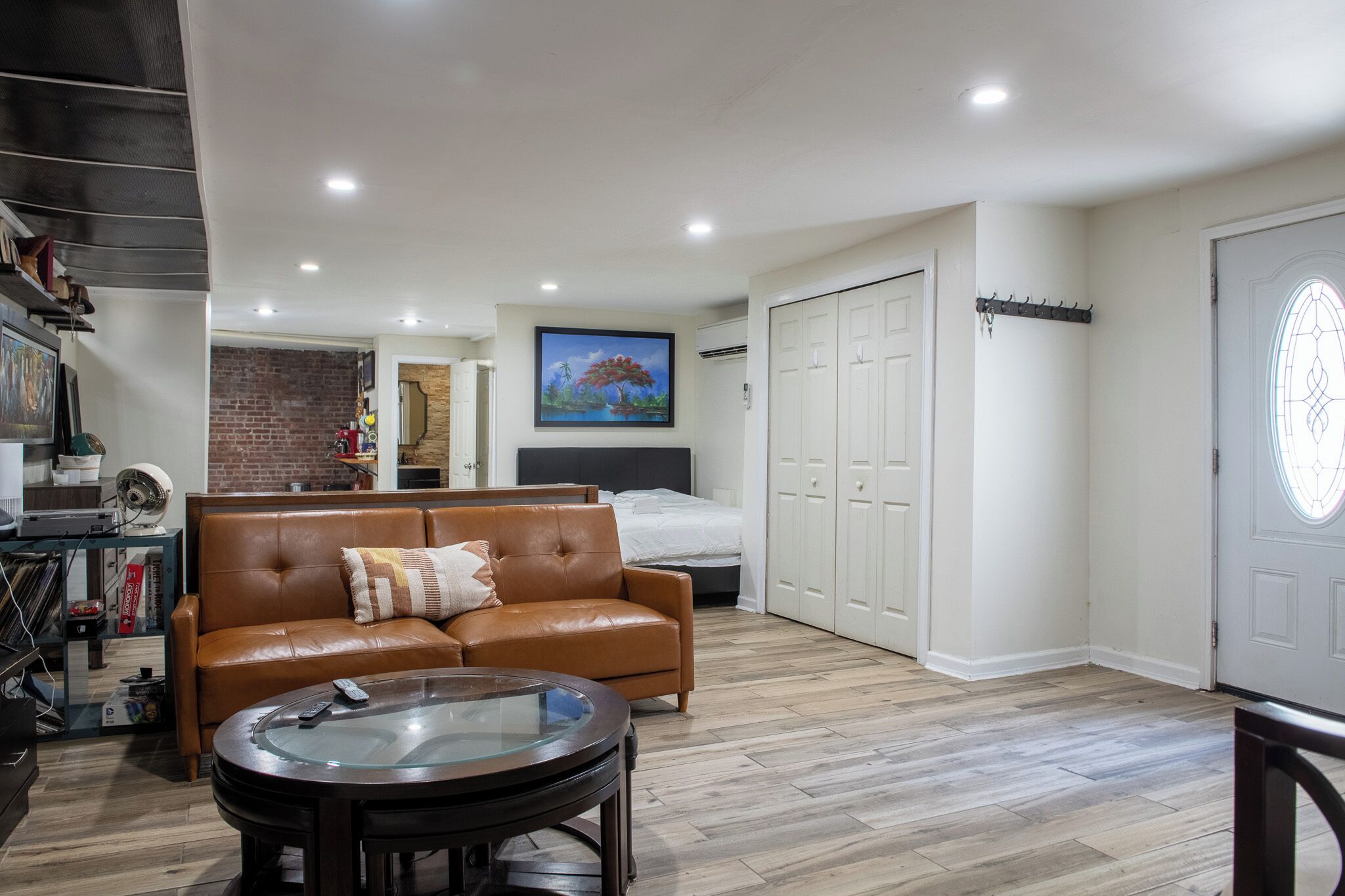 A modern living room with a leather sofa, a glass coffee table, and a bed with a headboard. There is a painting on the wall, a brick wall, and a white door with a coat rack.