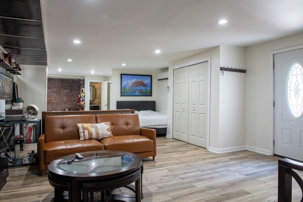 A modern living room with a leather sofa, a glass coffee table, and a bed with a headboard. There is a painting on the wall, a brick wall, and a white door with a coat rack. - Near NYC & Metro-North! Private Yonkers Studio (Yonkers)