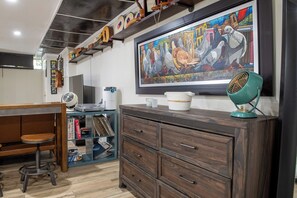 A room with a wooden desk, a large painting, a vintage fan, and a cabinet with drawers.