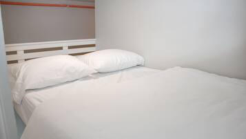 Una cama individual con ropa de cama blanca y almohadas, cabecera blanca y pared lisa.