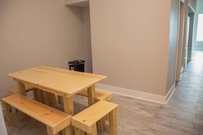A room with a wooden table and benches, a trash bin, and a hallway with a patterned floor.