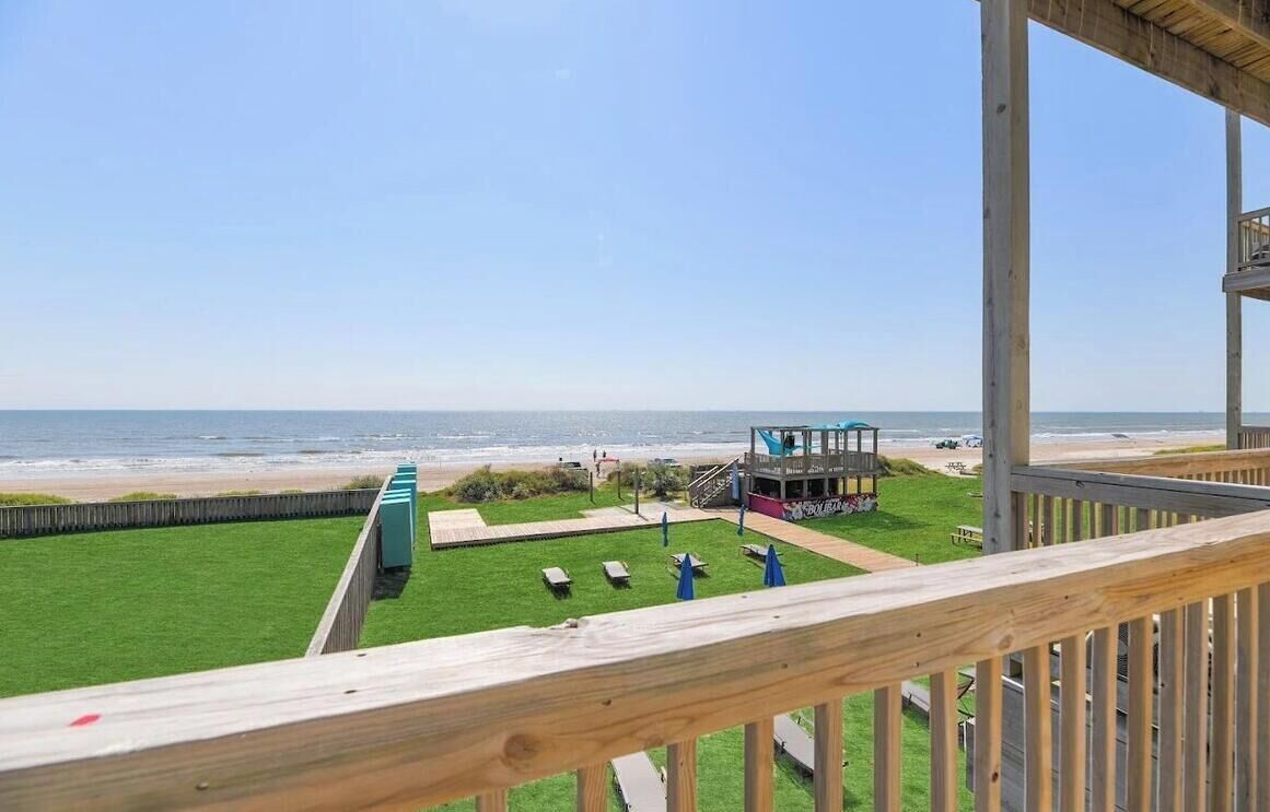 A beach view from a wooden deck with a lifeguard tower, green lawn, and beach umbrellas.