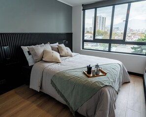 Quarto moderno com cama, almofadas, mesa de cabeceira e janela com vista para a cidade.