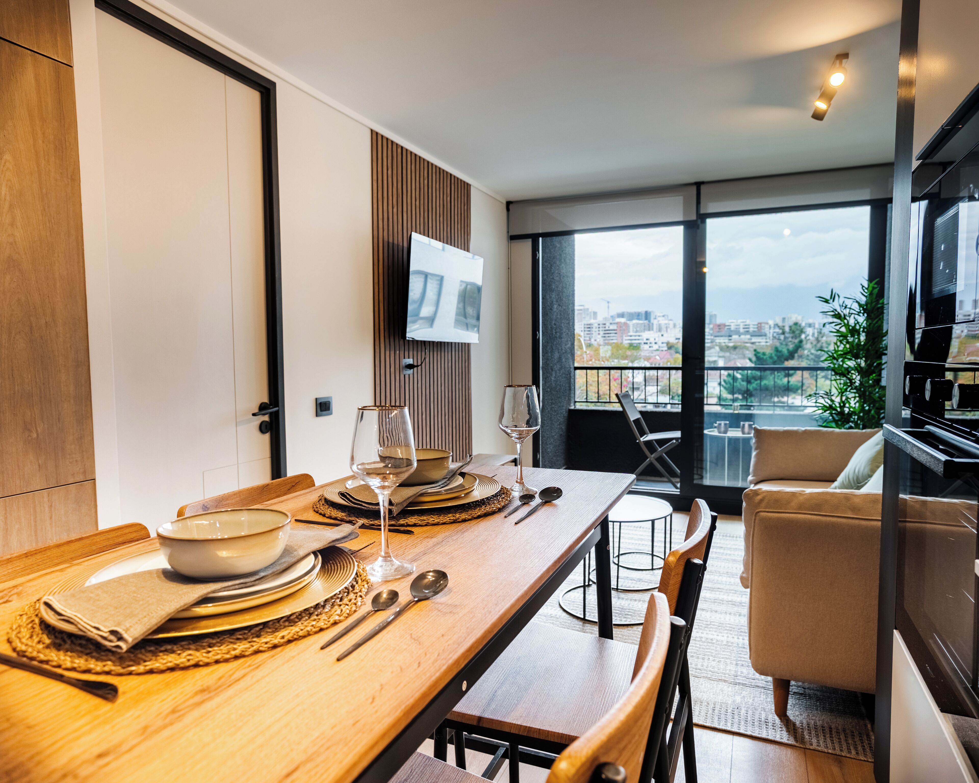 A modern dining area with a wooden table set for two, a television mounted on the wall, and a view of the city through large windows.