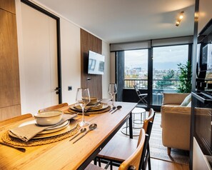A modern dining area with a wooden table set for two, a television mounted on the wall, and a view of the city through large windows. - San Eugenio Apartments by Andes STR (Ñuñoa)