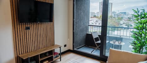 Una sala de estar moderna con televisor de pantalla plana, pared de paneles de madera, un sofá, dos mesitas y un balcón con vista a la ciudad.