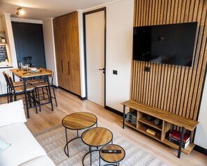 Uma sala de estar moderna com piso de madeira, área de jantar com mesa e cadeiras, televisão montada em uma parede de painéis de madeira e um banco com espaço de armazenamento por baixo.