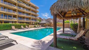 A hotel pool area with lounge chairs, a thatched-roof structure, and a multi-story building with balconies.
