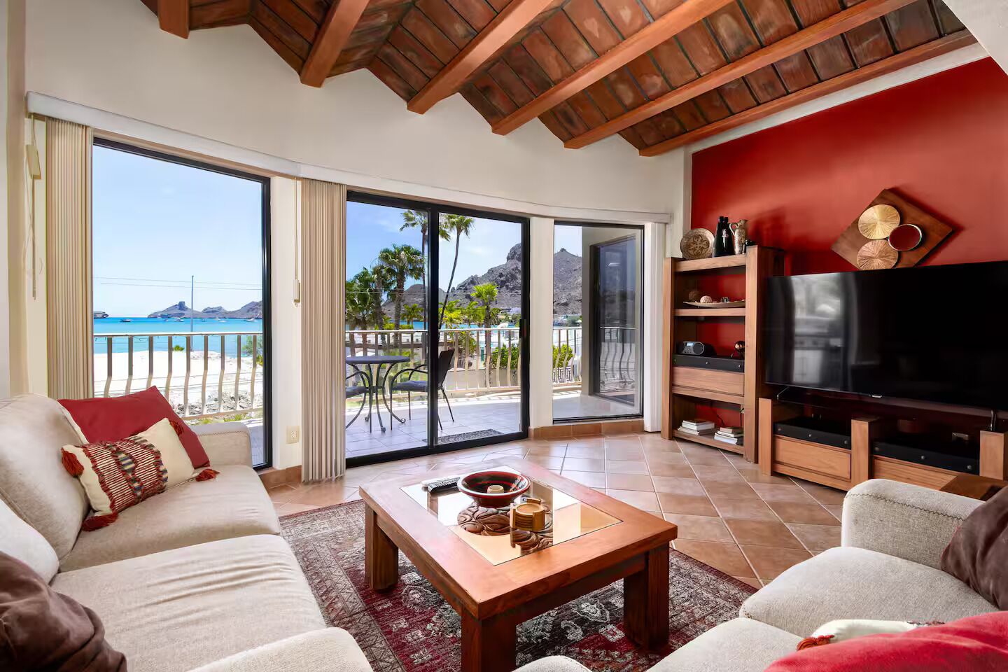 Un salon avec un canapé, une table basse et un téléviseur. Il y a un balcon avec vue sur la plage et les palmiers.