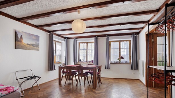 Una habitación con piso de madera, una mesa de comedor con sillas, una lámpara colgante y una ventana con cortinas.