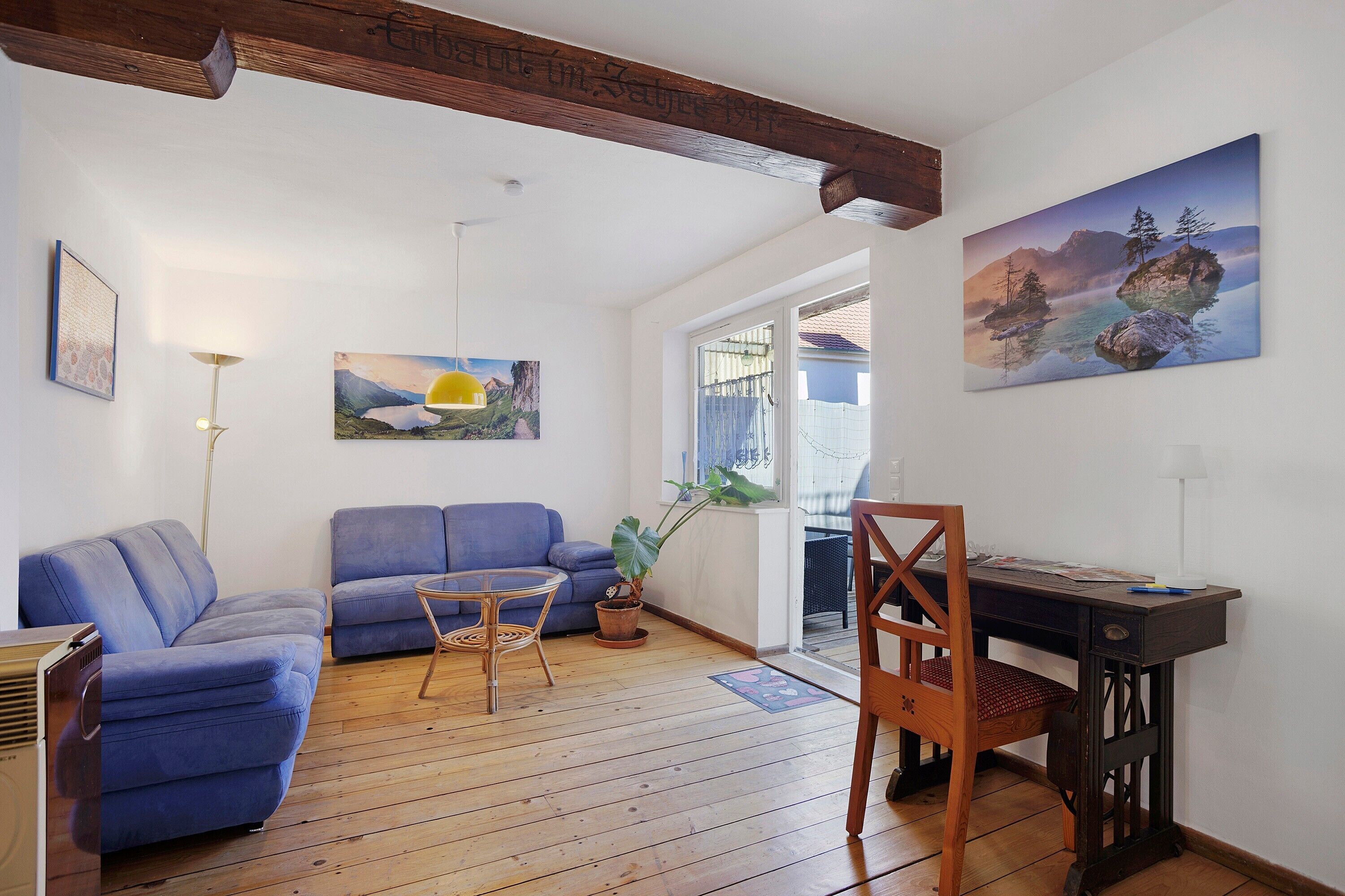 A living room with a blue sofa, a wooden dining table with a chair, and a painting of a mountain landscape.