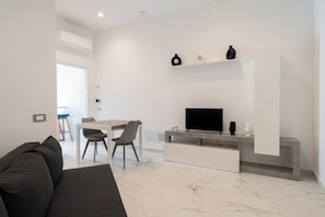 A modern living room with a sofa, a dining table with chairs, a flat-screen TV, and a minimalist shelf with decorative items. - The Urban Nest White & Gray (Bari)