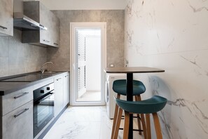 A modern kitchen with a built-in oven, a stainless steel range hood, and a dining area with a round table and two bar stools. - The Urban Nest White & Gray (Bari)
