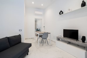 Een moderne woonkamer met een zwarte bank, een eettafel met stoelen, een flatscreen televisie en een plank met decoratieve objecten.