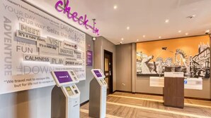 Zona de recepción de hotel con mostrador de registro, quiosco digital y una pared con frases motivacionales y un mural de la ciudad.