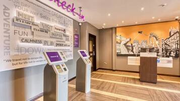 A hotel reception area with a 'check-in' desk, a digital kiosk, and a wall with motivational phrases and a cityscape mural.