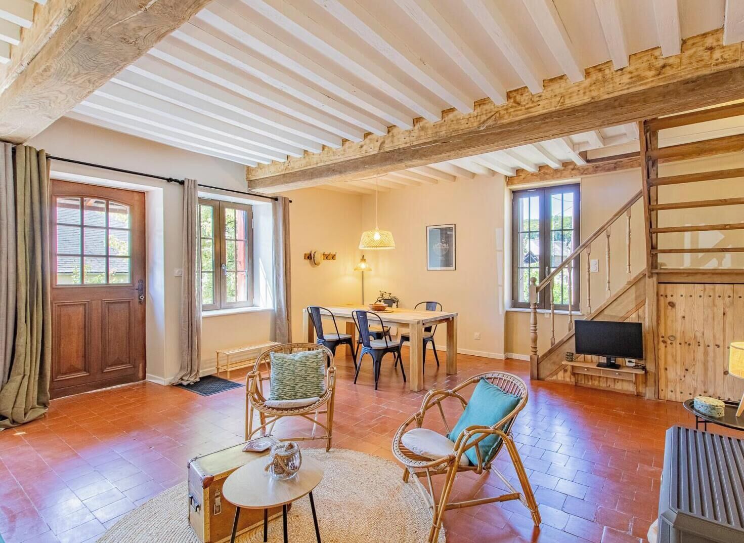 A spacious living area with a wooden ceiling, a dining table with chairs, a television, and a staircase.