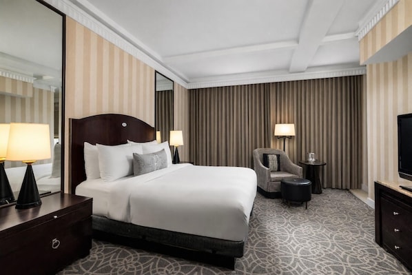 A hotel room with a large bed, a chair, a small table, a TV, and a mirror.