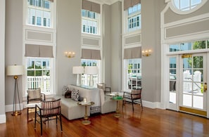 A spacious living room with a high ceiling, large windows, and a view of the outside. There are multiple seating areas, including a sofa and chairs, and a coffee table.