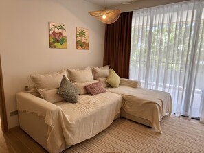 Ein beiges Ecksofa mit gemusterten Kissen, eine gewebte Pendelleuchte und zwei Wandbilder mit tropischen Motiven.