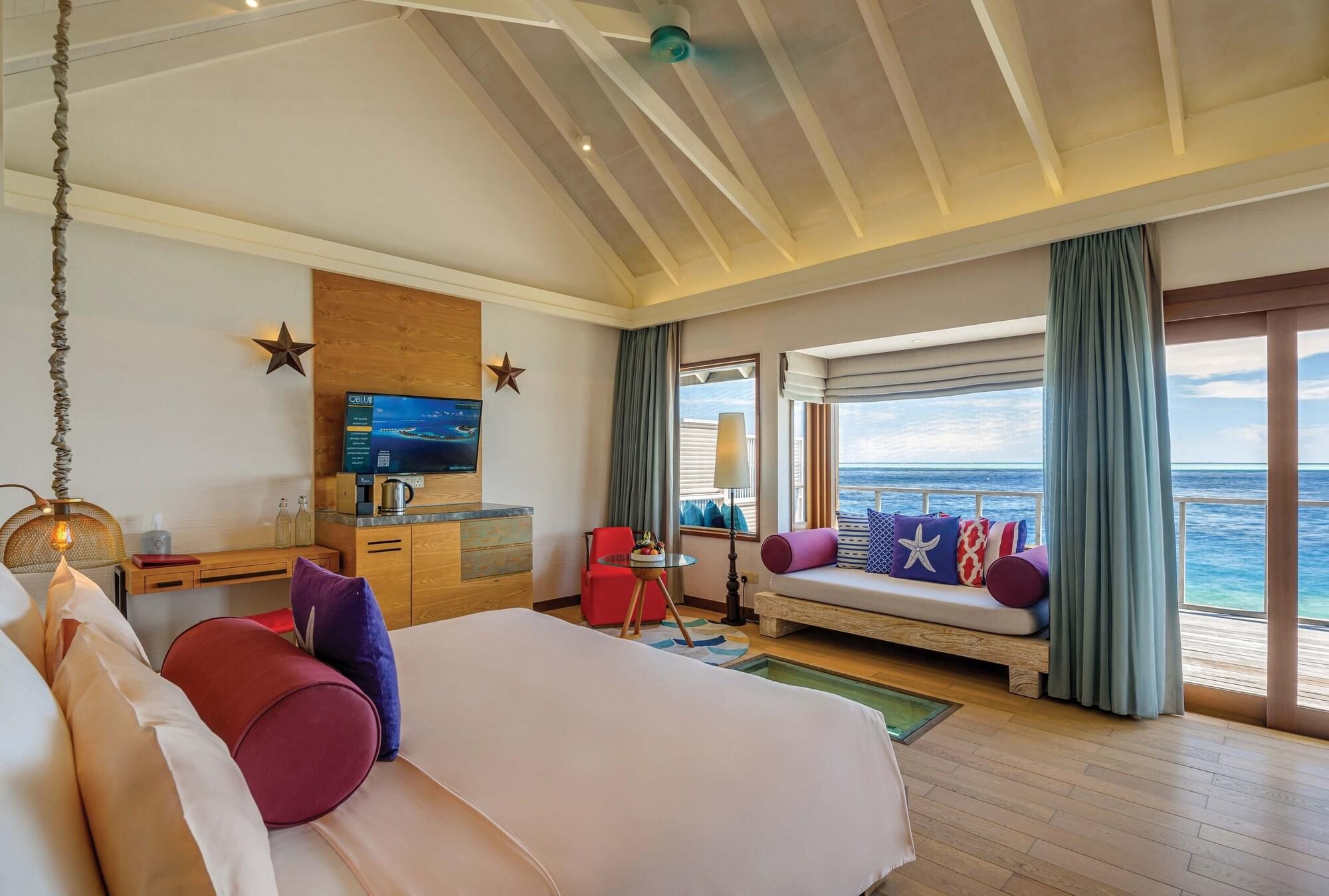 A hotel room with a bed, a desk with a coffee maker, a TV, a seating area with cushions, and a view of the ocean.