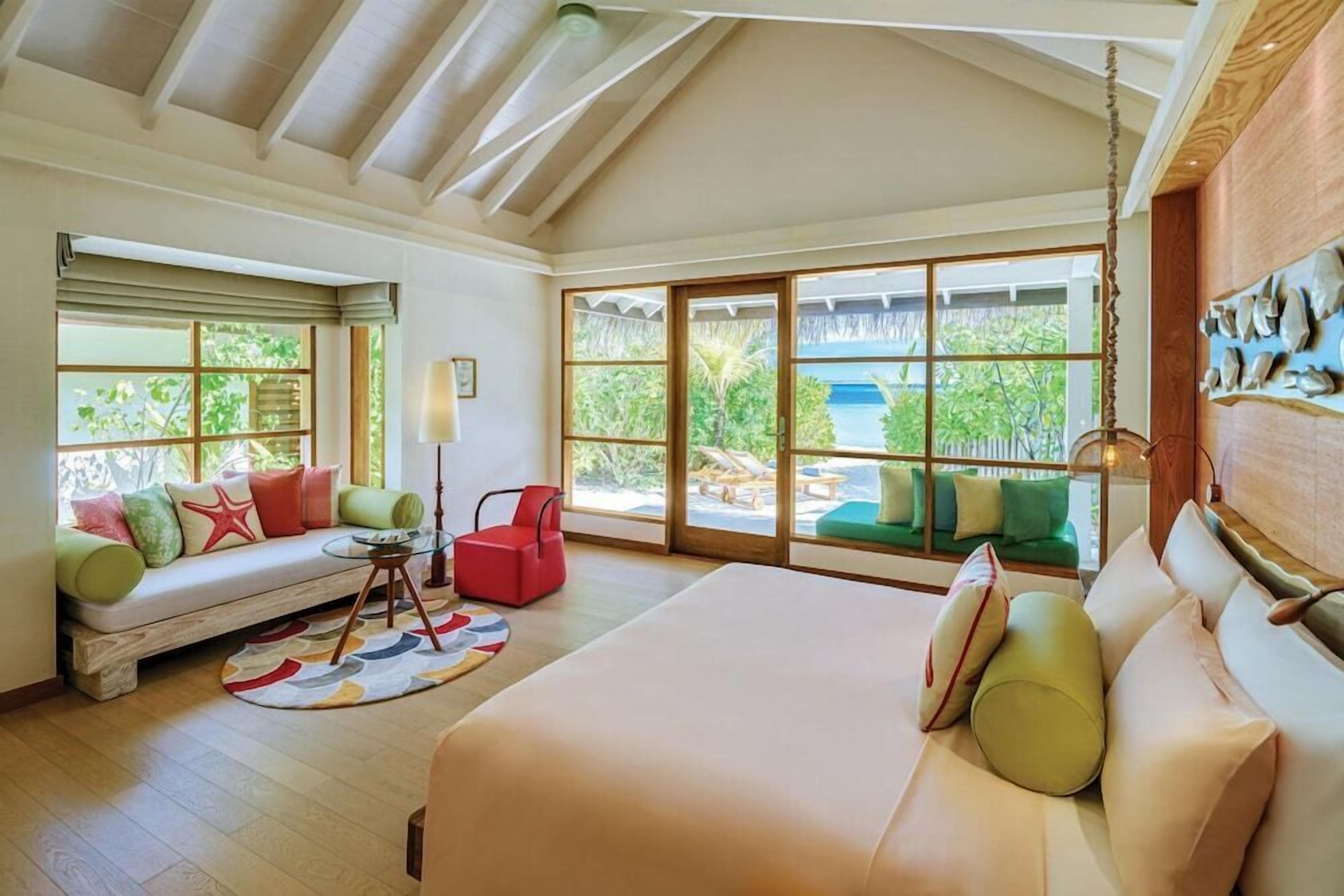 Ein geräumiges Zimmer mit einem großen Bett, einer Couch und einem roten Sessel. Große Fenster bieten Blick ins Freie.