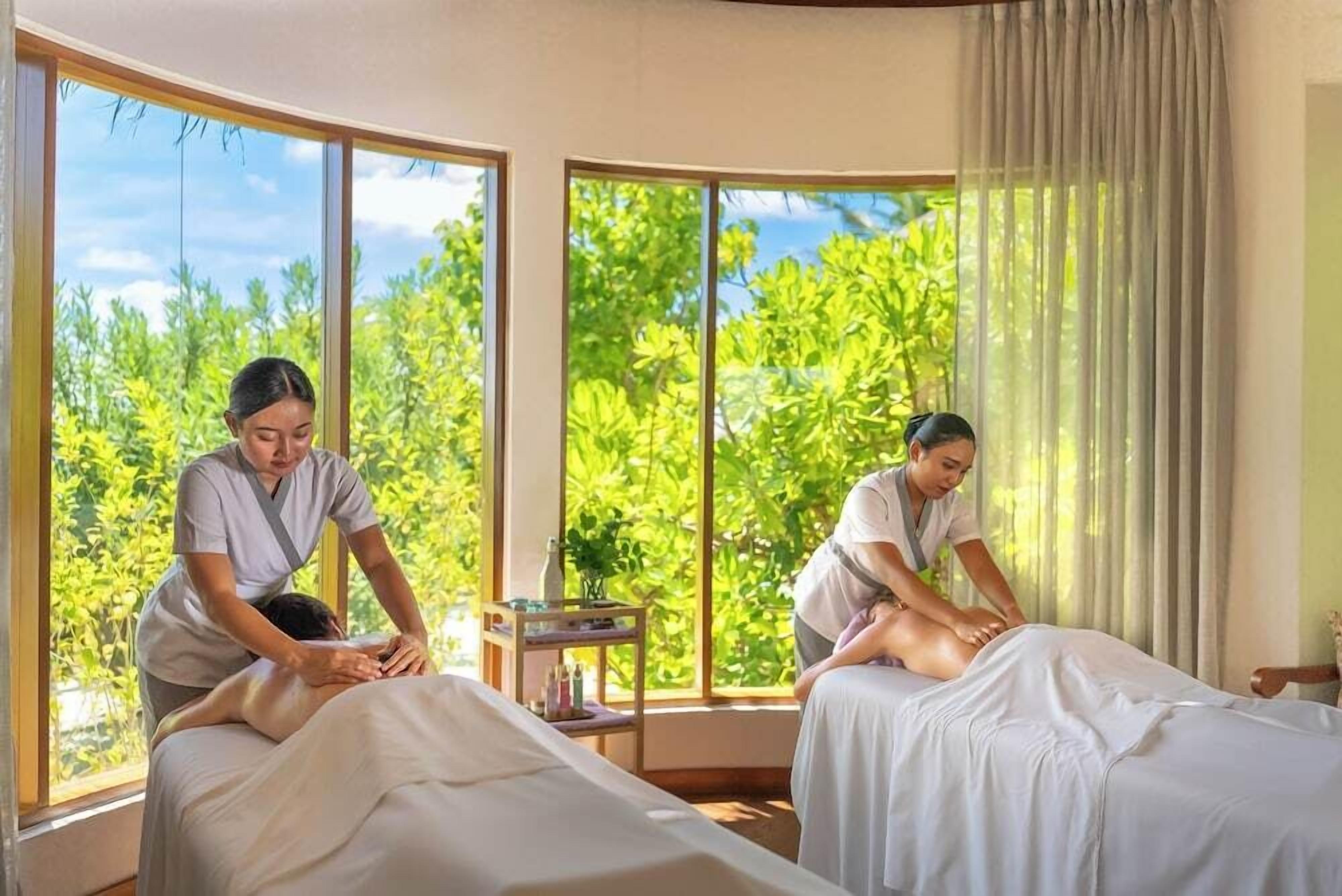 A room with two beds, large windows, and a view of greenery outside.