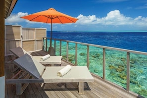 A wooden deck with lounge chairs and an umbrella, overlooking clear blue water and a tropical beach.