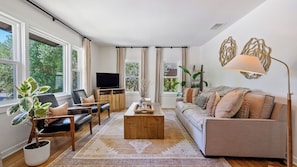 A modern living room with a sofa, two armchairs, a coffee table, and a television. There are large windows with curtains, a potted plant, and a floor lamp. - Lavender Porch by Avantstay Designer Bungalow + Fire Pit Near Downtown, Paso (Paso Robles)