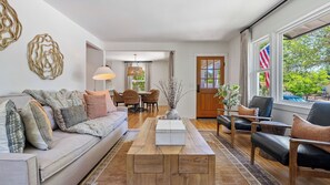 Uma sala de estar moderna com sofá, mesa de centro e cadeiras, área de jantar com mesa e cadeiras, e janelas amplas com uma bandeira americana.