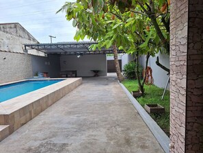 Ein Schwimmbecken mit überdachter Terrasse, einem Betonweg und einem kleinen Garten mit einer Palme.