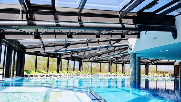En inomhuspool med ett tak av genomskinligt glas, gröna solstolar och utsikt över grönområden utanför.