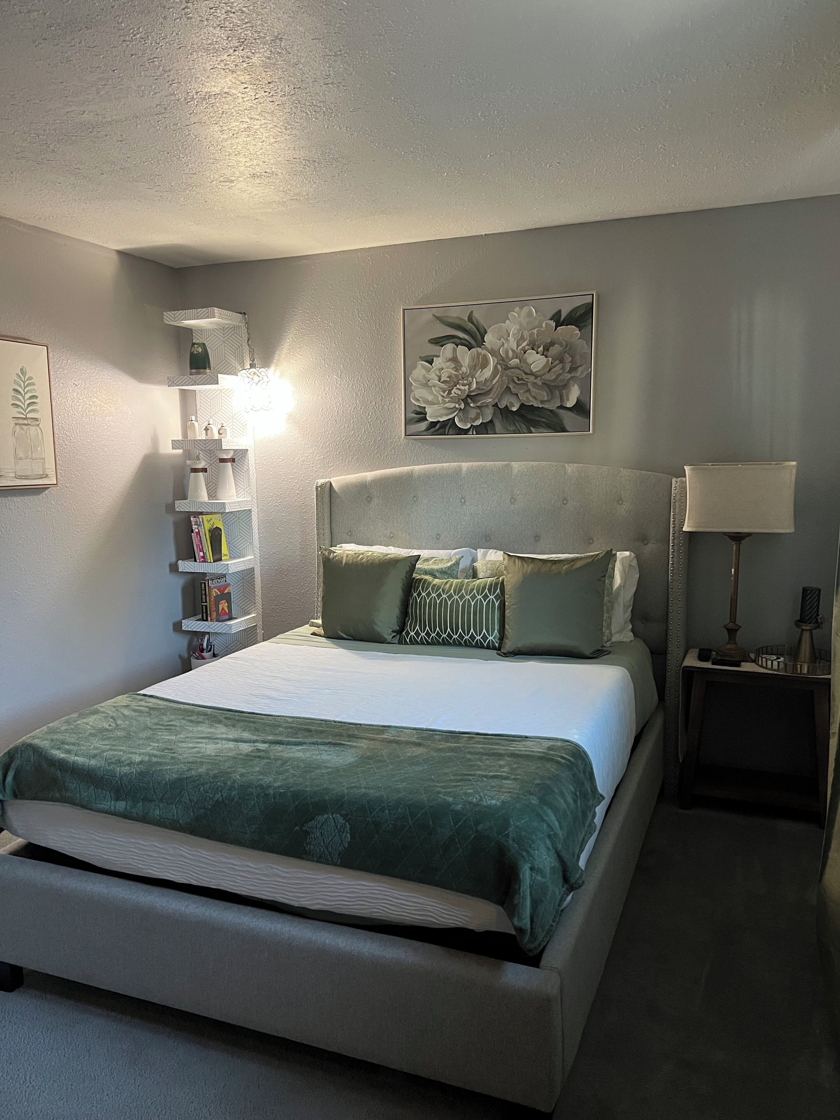 Une chambre à coucher bien rangée, avec un lit, des tables de chevet, une lampe et une étagère fixée au mur, où sont posés des livres et des objets décoratifs.