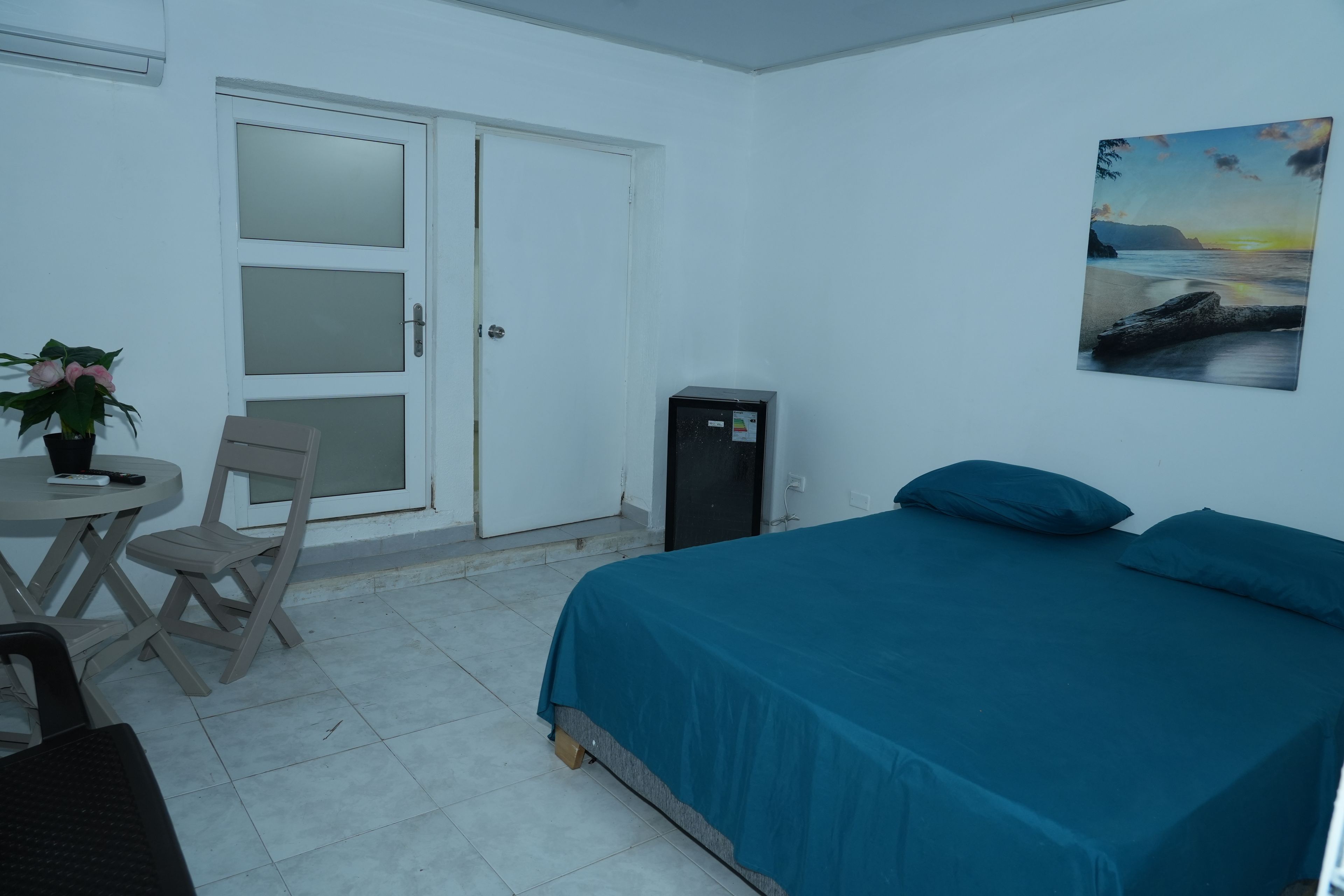 Quarto com cama, escrivaninha e cadeira, pequena mesa com planta e porta com painéis de vidro jateado.