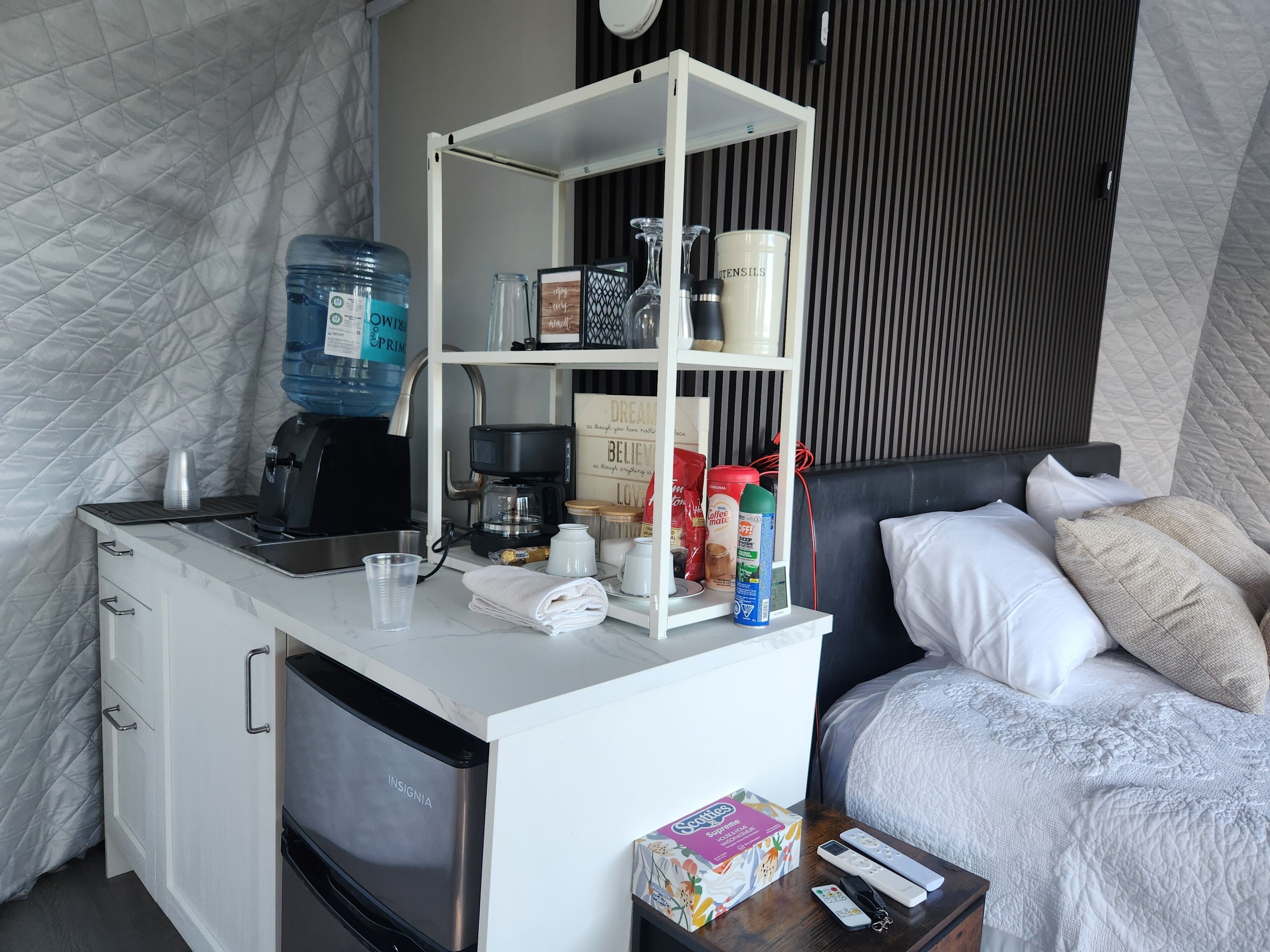 Una kitchenette compacta con cafetera, dispensador de agua y estantes para guardar objetos.