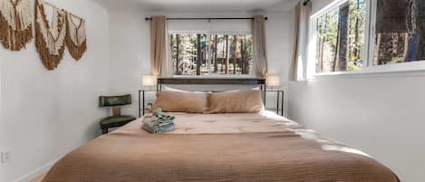 A bedroom with a large bed, a window with a view of trees, and a wall-mounted macrame piece.