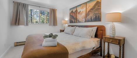 A bedroom with a large bed, a window with curtains, a painting above the bed, and a bedside table with a lamp.