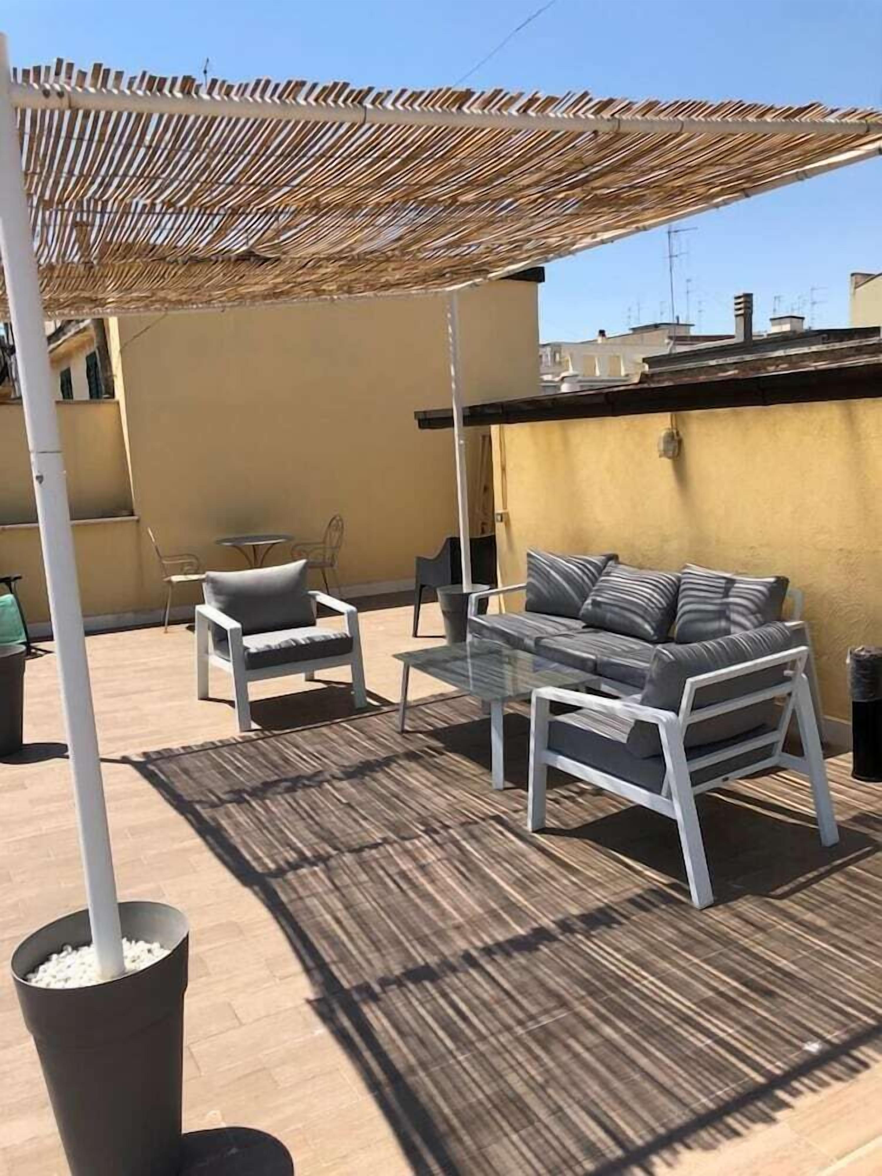 Een terras buiten met een tafel en stoelen, een geweven parasol en een bloempot.